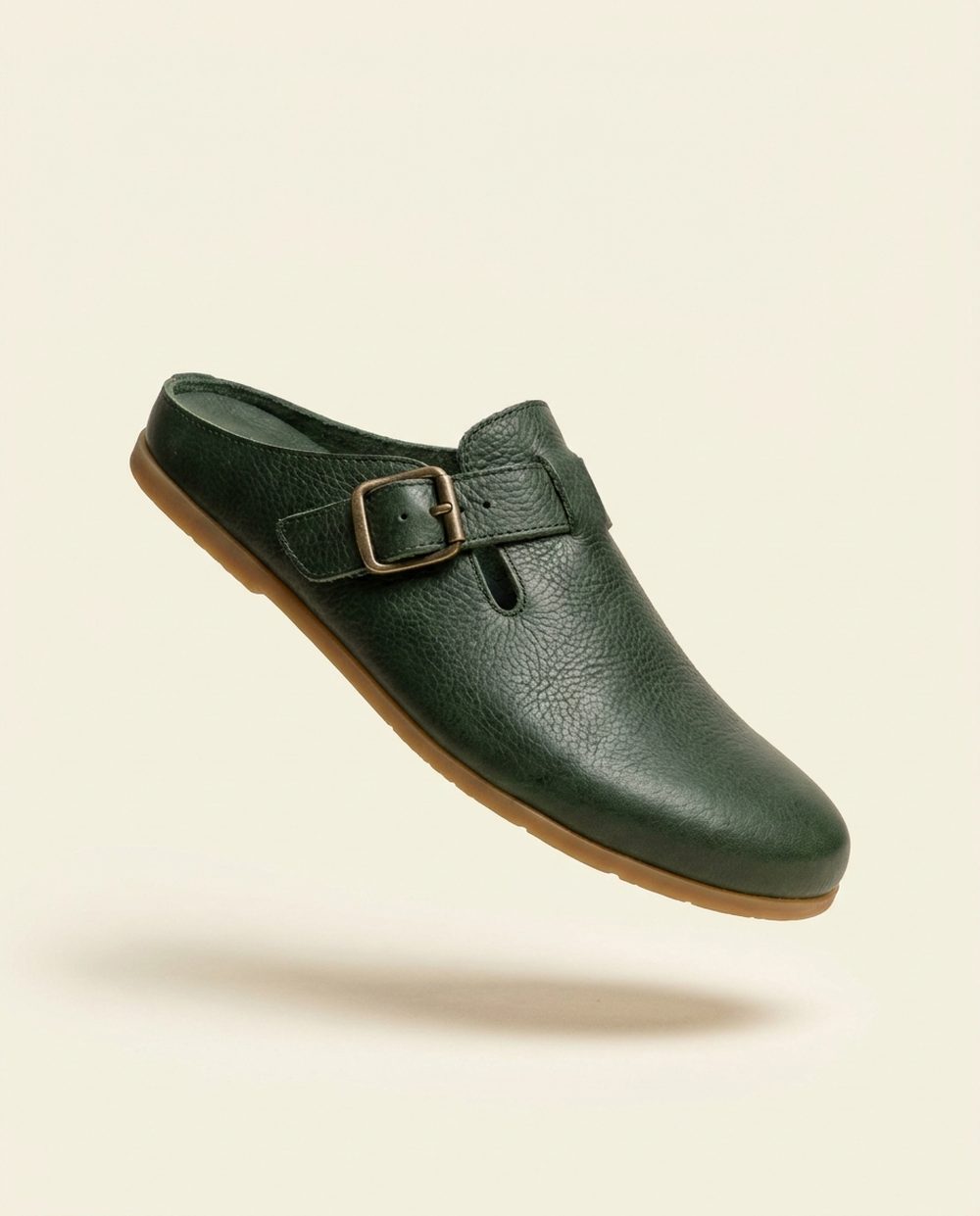 A minimalist barefoot mule in deep forest green pebbled floater leather with an antique brass buckle strap and a thin gum rubber sole